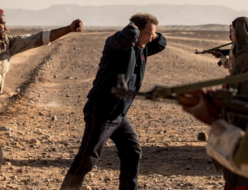 Dans le désert, un homme (Vincent Lindon) a les bras derrière la tête. Des armes sont pointées sur lui.