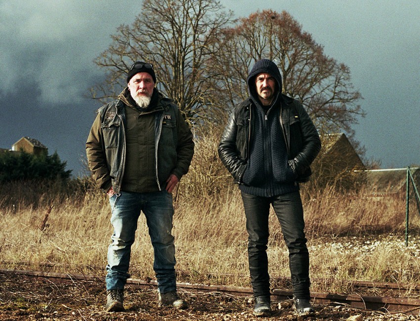 Deux hommes sont debout, l'un avec une capuche, l'autre avec un bonnet. Ils sont dans un endroit rural et désert.