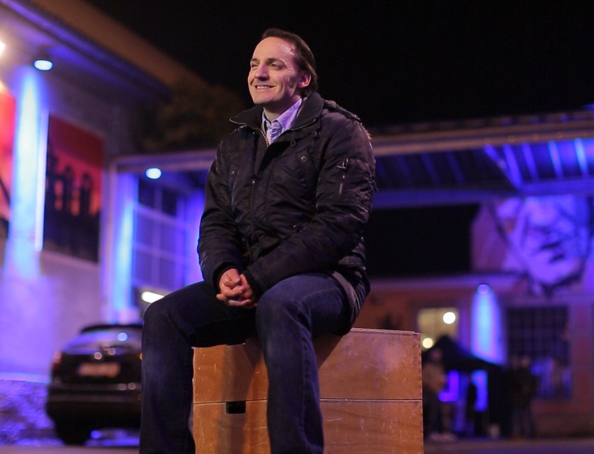 Un homme (Fabrizio Rongione) est assis sur une boite carrée. Il y a des lumières bleues derrière lui.