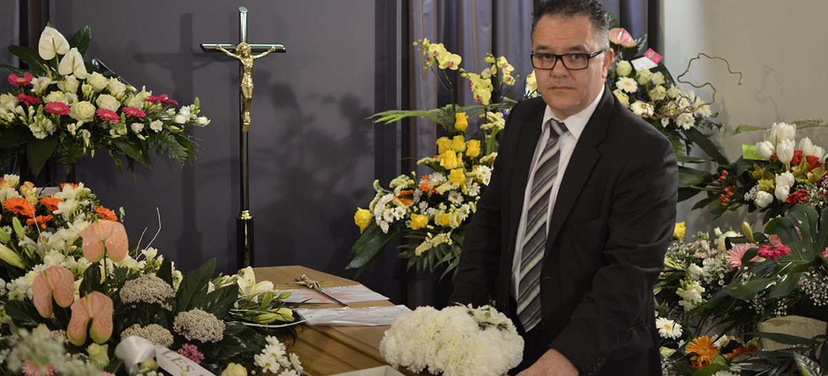 Un croque-mort s'appuie sur un cercueil, devant un crucifix et au milieu de fleurs multicolores.