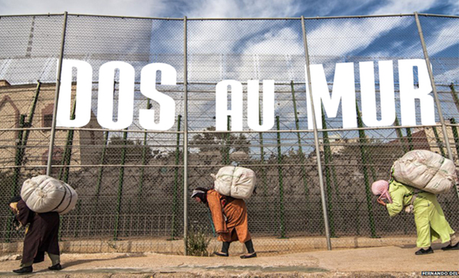 Des personnes portent un ballot sur leur dos et courbent l'échine, devant un grillage.