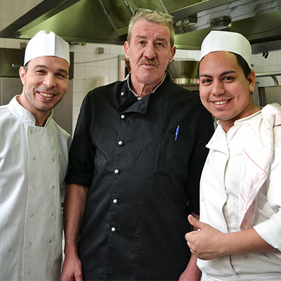 Un chef en tenue de cuisine noire pose à côté de deux commis en formation.