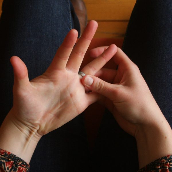 Delhie joue avec sa bague de fiançaille nerveusement, la photo est prise en plongée. On ne voit que ses genoux et ses mains.