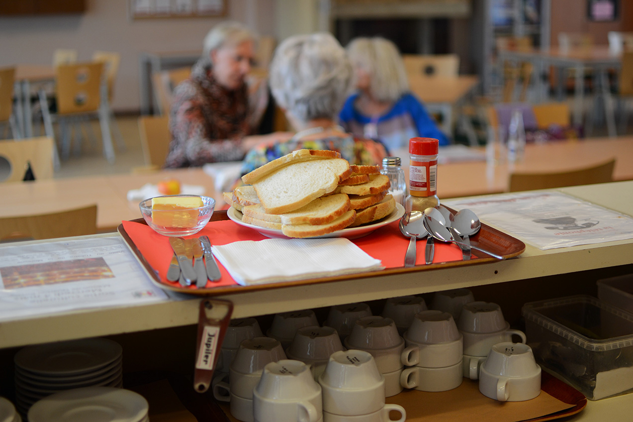 Sur un bar, un plateau est rempli de tartines et de cuillères. Au fond, trois dames agées sont installées à une table. 