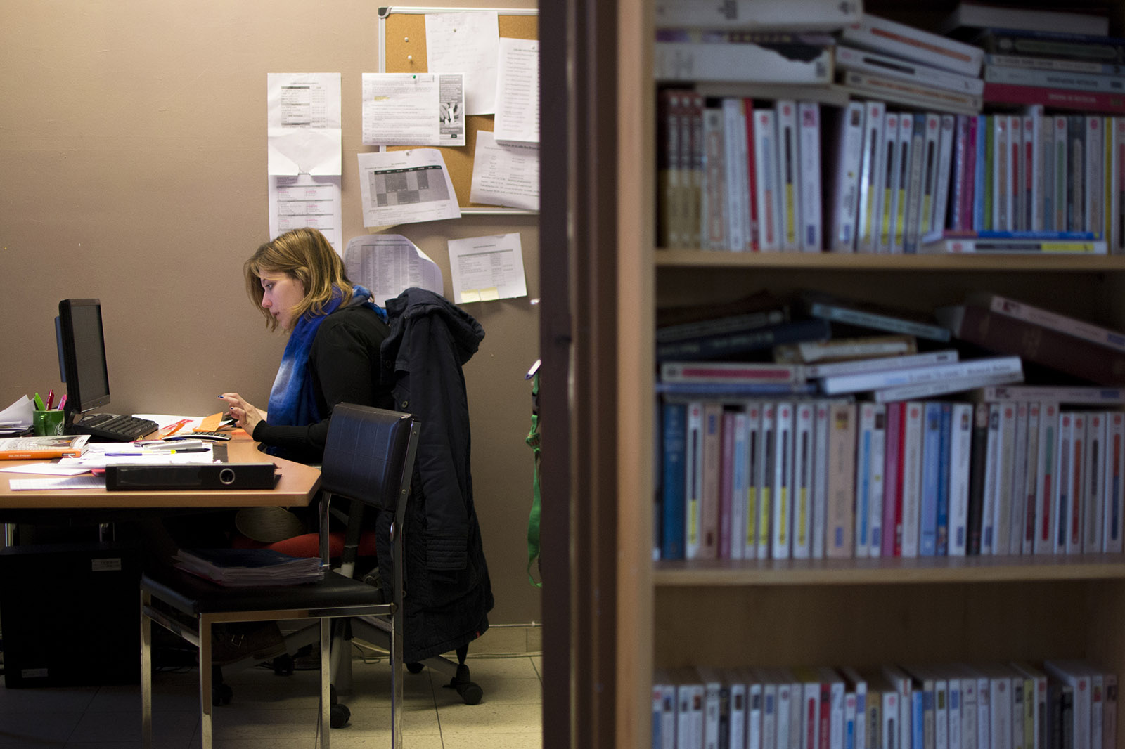 A gauche, une jeune femme est installée à son bureau, devant un ordinateur. A droite se trouve une bibliothèque remplie de livres.